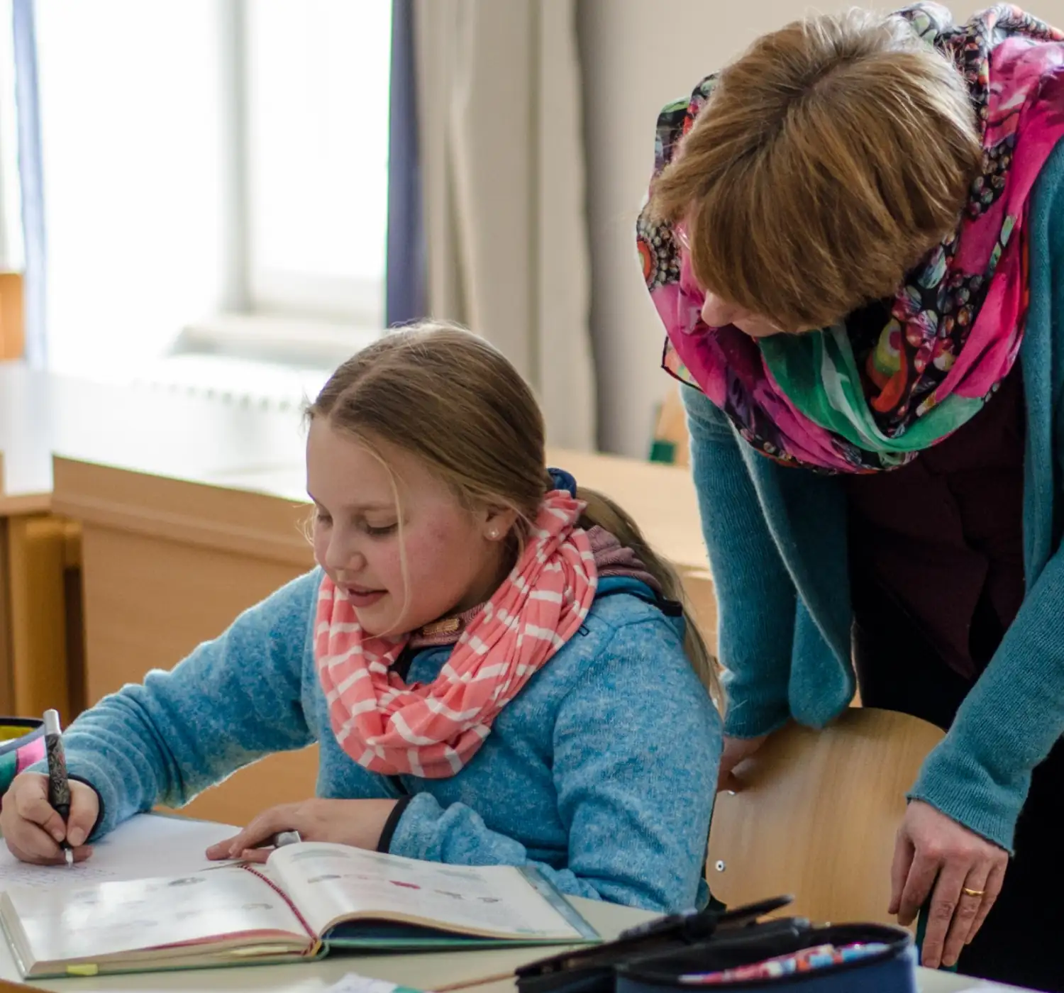 Eine Schülerin beim Lernen mit einer Lehrerin im Klassenzimmer. Beide tragen warme Schals und konzentrieren sich auf das Buch.