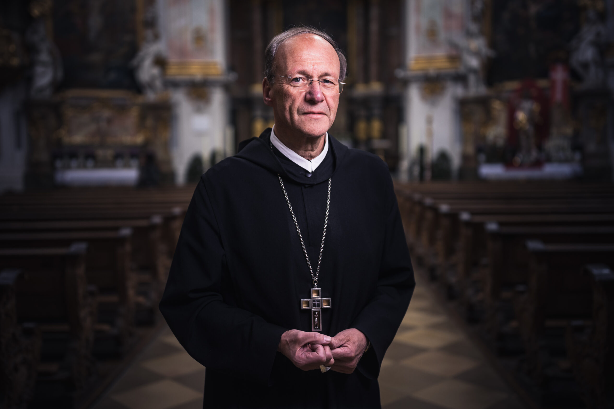 Schwangerer Priester in der Kirche mit Kreuz um den Hals Ein Priester in einem Kircheninneren trägt ein großes Kreuz und steht inmitten leerer Bankreihen.
